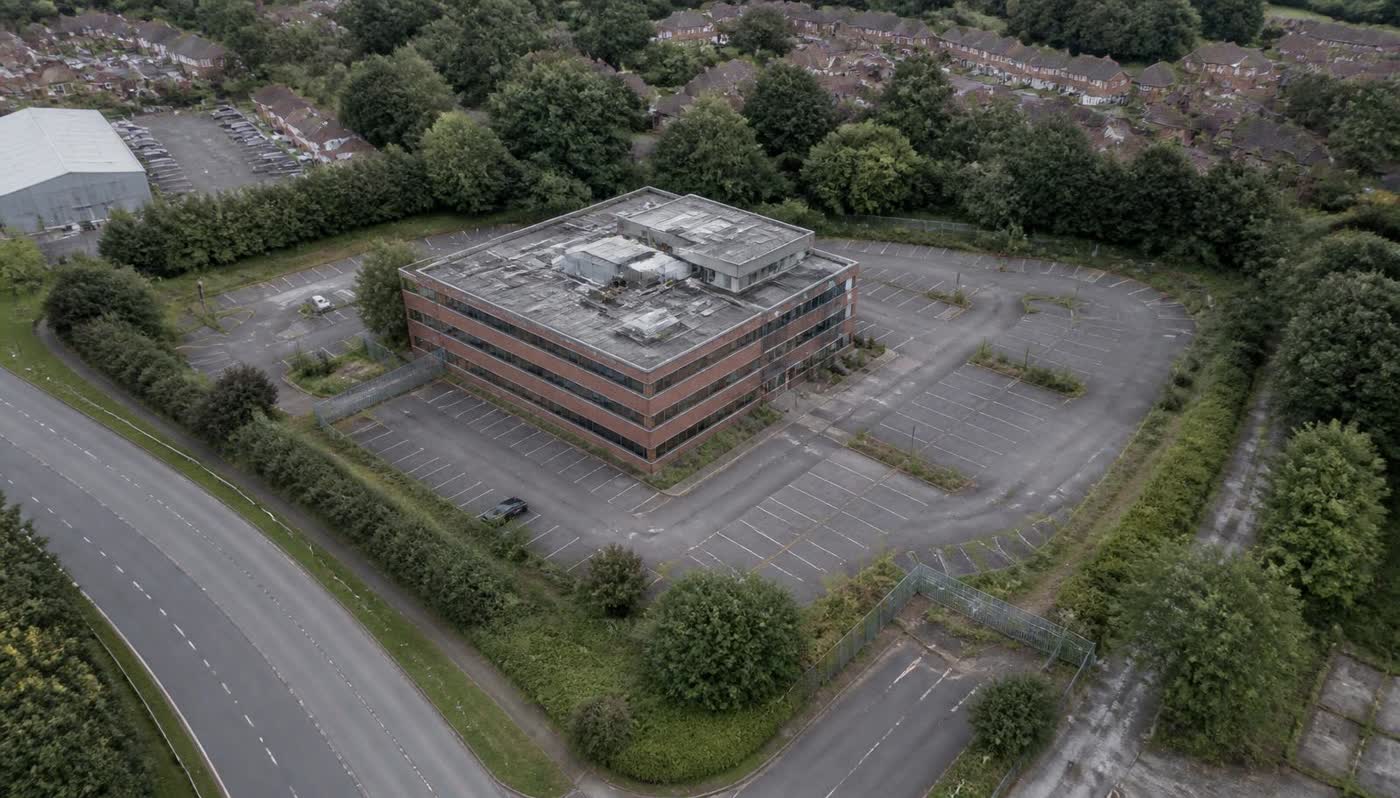 Aerial oblique view of a dated three-storey suburban office building with a large half-empty car park, overcast light